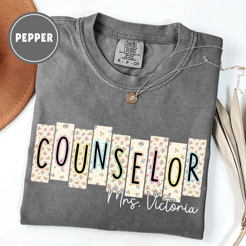 May include: A gray Comfort Colors t-shirt with the word "COUNSELOR" in a floral design, and the name "Mrs. Victoria" below. A silver necklace with a pendant is draped on the shirt.