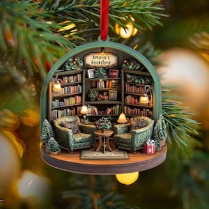 May include: A round, hanging Christmas ornament featuring a detailed miniature bookstore interior with two green patterned armchairs, a small center table, and bookshelves filled with books. A sign above reads Amelia's Bookstore.