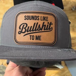 May include: Gray trucker hat with a brown leather patch. The patch features the phrase "SOUNDS LIKE Bullshit TO ME" in a bold, stylized font. The hat has a mesh back and a curved brim.