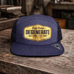 May include: Navy blue flat-brimmed cap with a black mesh back. The cap features a gold and black patch that reads "Blue Collar Degenerate" with additional text. The cap is sitting on a wooden surface.