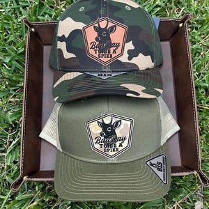 May include: Three camouflage and olive green trucker hats with the text "Bad Day To Be A Spike" on a patch featuring a deer silhouette. The hats are displayed in a brown leather tray on a bed of green grass.