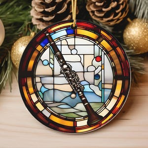 May include: Round Christmas ornament featuring a stained glass design with an oboe in the center. The ornament has a gold string for hanging. The background includes pine needles, pine cones, and gold and silver ornaments.