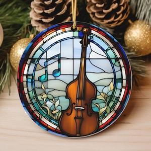 May include: Round Christmas ornament with a stained glass design featuring a brown upright bass and musical notes. The ornament has a gold string for hanging and is surrounded by pine needles, pine cones, and gold and silver ornaments.