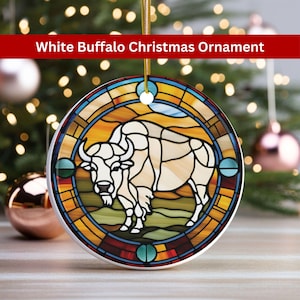 May include: A white ceramic Christmas ornament featuring a stained glass design of a white buffalo in a circular frame with yellow, orange, red, blue, and green accents.