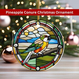 May include: Round Christmas ornament with a stained-glass design featuring a colorful pineapple conure parrot perched on a branch. The ornament has a gold string for hanging and the text "Pineapple Conure Christmas Ornament" in a red banner.