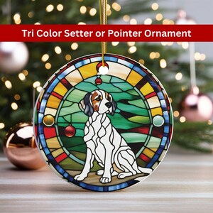 May include: A round ceramic ornament featuring a stained glass design of a tri-color setter or pointer dog sitting in a green field. The ornament has a gold string for hanging.