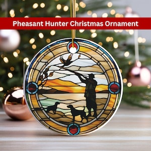 May include: A round ceramic Christmas ornament featuring a stained glass design of a hunter with two dogs, a pheasant in flight, and a sunset landscape. The ornament has a gold ribbon for hanging.