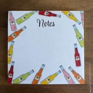 May include: A square notepad with a light blue background and a border of colorful beverage bottle illustrations. The word "Notes" is written in black script in the center of the notepad.