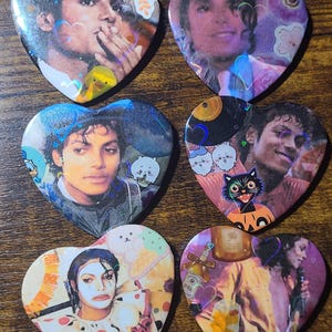 May include: Six heart-shaped pins featuring various photographic portraits of Michael Jackson. The designs include holographic heart overlays, cartoon illustrations of animals, food, and festive items, with a glossy finish on each individual pin.
