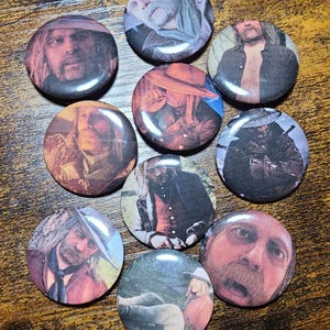 Könnte beinhalten: Eine Sammlung von neun runden Ansteckbuttons mit Porträts von Filmfiguren. Die Buttons zeigen verschiedene Schauspieler in Kostümen mit unterschiedlichen Gesichtsausdrücken und Outfits. Die Buttons sind auf einer Holzoberfläche angeordnet.