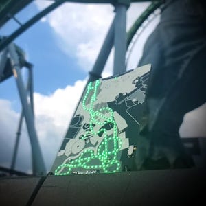 May include: A rectangular sign with a map design and green neon lights. The sign has the text "LIGHTRIDERS" at the bottom. The background shows a blue sky and a roller coaster structure.
