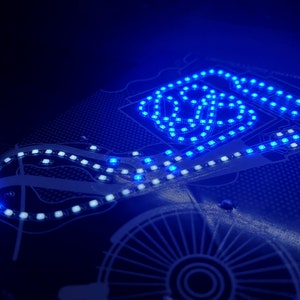May include: A black and white printed circuit board with a blue LED light strip outlining a race track design.