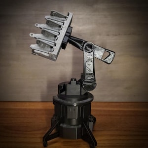 May include: A 3D-printed model of a mechanical device, featuring a gray, articulated arm and a multi-pronged head. The base is dark gray, with a tripod-like structure. The arm and head are gray and black, with intricate details.