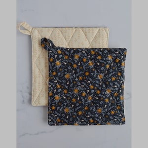 May include: Two quilted pot holders. One is cream-colored with a subtle leaf pattern, and the other is navy blue with a floral design in yellow and white. Both have a loop for hanging and a diamond-stitched pattern.