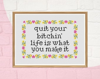 May include: Framed cross-stitch artwork with the text "quit your bitchin' life is what you make it". The black text is set against a white background, framed by a floral border with yellow and pink flowers.