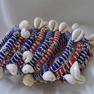 May include: A collection of colorful bracelets. Each bracelet is wrapped in red and blue fabric with silver accents. White seashells are attached to the bracelets. The bracelets are arranged on a white surface.