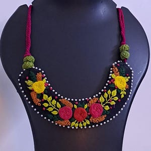 Peut inclure: Un collier brodé noir et un ensemble de boucles d'oreilles assorties. Le motif floral présente des fleurs jaunes, rouges et roses avec des feuilles vertes, soulignées de perles blanches. Le collier est présenté sur un mannequin noir.