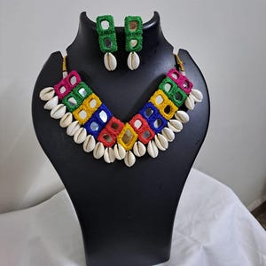 May include: A colorful necklace and earring set displayed on a black mannequin. The necklace features a vibrant array of squares in red, pink, yellow, green, and blue, adorned with small mirrors and white seashells. The matching earrings are green squares with seashells.