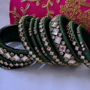 May include: A collection of green bangles with square and round embellishments. The bangles are arranged in a row, with a pink embroidered clutch in the background. A small jar with gold beads is also visible.