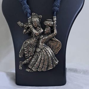 Può includere: Collana con pendente argentato e orecchini abbinati. Il pendente raffigura due figure in abiti tradizionali, con un flauto e un vaso. La collana ha un cordino blu intrecciato. Gli orecchini hanno un design abbinato.