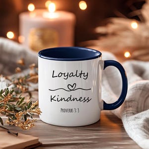 May include: A white ceramic mug with a dark blue interior and handle. The front features the black text "Loyalty" above a small heart icon and a wavy line, followed by the word "Kindness" and the citation "Proverbs 3:3" in black font.