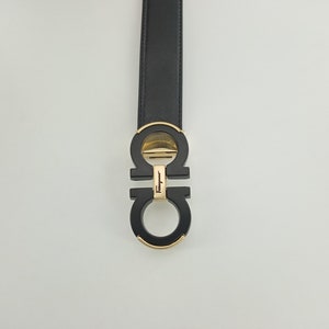 以下が含まれることがあります： ゴールドトーンのバックルが付いた黒いベルト。バックルはブランド名入りのダブルGデザインです。ベルトは質感のある素材でできており、白い背景に配置されています。