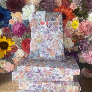 Puede incluir: Tres cajas de regalo envueltas en papel con estampado floral con flores moradas, naranjas y blancas. Un cuarto regalo est&aacute; envuelto en el mismo papel y cubierto con pegatinas de coraz&oacute;n y una pegatina de mariposa. El fondo es una pared de flores coloridas.