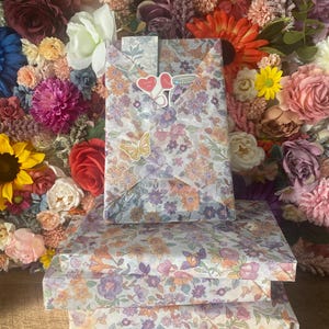 Puede incluir: Tres cajas de regalo envueltas en papel con estampado floral con flores moradas, naranjas y amarillas. La caja superior est&aacute; decorada con pegatinas de coraz&oacute;n y tarro. El fondo es una pared de flores coloridas.