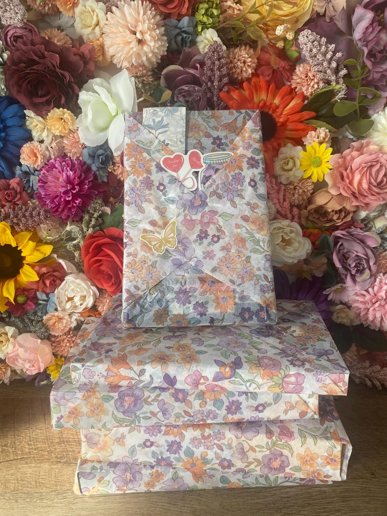 Puede incluir: Tres cajas de regalo envueltas en papel con estampado floral con flores moradas, naranjas y amarillas. La caja superior est&aacute; decorada con pegatinas de coraz&oacute;n y tarro. El fondo es una pared de flores coloridas.
