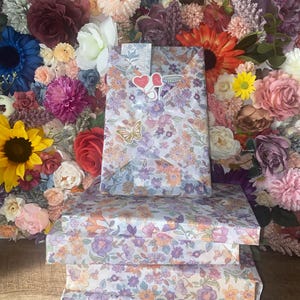 Puede incluir: Tres cajas de regalo envueltas en papel con estampado floral con flores moradas, naranjas y blancas. Una bolsa de regalo a juego est&aacute; encima, adornada con pegatinas de coraz&oacute;n y una pegatina de mariposa. El fondo es una pared de flores coloridas.