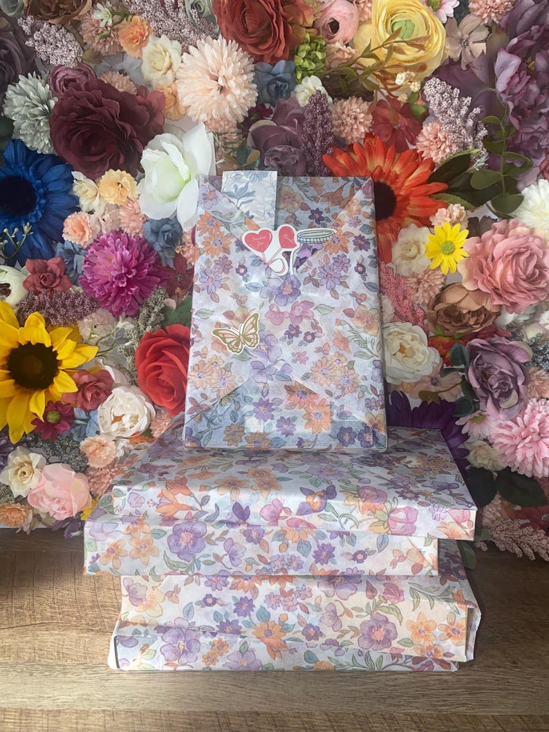 Puede incluir: Tres cajas de regalo envueltas en papel con estampado floral con flores moradas, naranjas y blancas. Una bolsa de regalo a juego est&aacute; encima, adornada con pegatinas de coraz&oacute;n y una pegatina de mariposa. El fondo es una pared de flores coloridas.