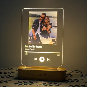 May include: A white and gold acrylic music player with a photo of a couple in a car. The player has a black and white progress bar with the song title "You Are The Reason" by Calum Scott. The base of the player is made of wood and has the text "Just to be with you" engraved on it.