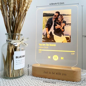 May include: A wooden music player night light with a clear acrylic panel displaying a photo of a couple and the song title "You Are The Reason" by Calum Scott. The base of the night light is engraved with the text "Just to be with you". The night light measures 19 cm tall, 14 cm wide, and 4 cm deep.