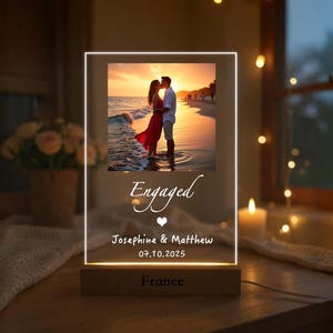 Pode incluir: Um expositor de fotos em acrílico iluminado com uma base de madeira. O expositor apresenta uma foto de um casal numa praia ao pôr do sol, com a palavra "Engaged" e nomes e data abaixo. A base diz "France".