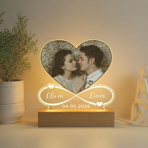 May include: A personalized heart-shaped LED lamp featuring a couple's photo. The lamp includes the names "Olivia" and "Liam" with the date "04.05.2025" displayed below. The base is made of wood, and the lamp emits a warm glow.
