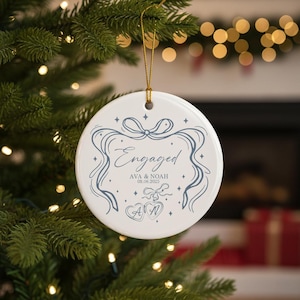 Può includere: Ornamento natalizio rotondo in ceramica bianca con cordino dorato. Presenta la scritta "Engaged" in blu-grigio, i nomi "Ava & Noah" e la data "08.08.2023". Decorato con nastri e cuori.