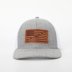 May include: A gray trucker hat with a white mesh back and a curved brim. The front features a brown leather patch with an American flag design and an airplane silhouette.