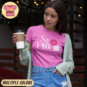 Peut inclure: T-shirt rose avec l'inscription "So Fetch" et une bouche rouge. Le t-shirt est porté avec un jean bleu et un cardigan vert clair. Une tasse de café est tenue à la main. Le t-shirt fait partie de la collection "Viral Style".