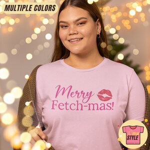 May include: A light pink t-shirt with the text "Merry Fetch-mas!" in pink script and a pink lip print. The shirt has a crew neck and short sleeves. The words "Multiple Colors" are at the top.