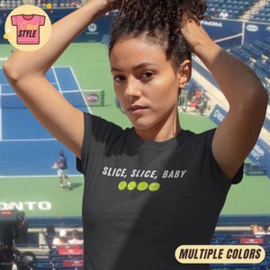 Puede incluir: Camiseta negra con la frase "SLICE, SLICE, BABY" y cuatro pelotas de tenis. La persona lleva la camiseta delante de una cancha de tenis. Las palabras "MULTIPLE COLORS" están en un cuadro amarillo.