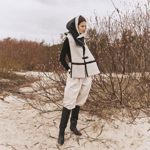 May include: A woman wearing a white and black hooded vest with a black turtleneck sweater, white pants, and black boots. She is standing in a field of brown branches.