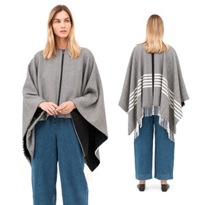 May include: A gray and black striped poncho with a zipper closure and fringe trim. The poncho is worn over blue jeans.