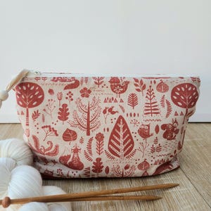 Peut inclure: Une pochette rectangulaire zippée en tissu beige clair avec un motif répétitif d'éléments forestiers rouges, incluant des arbres, des feuilles, des fleurs, un hibou, un écureuil et un renard. Une fermeture éclair blanche avec une perle en bois.