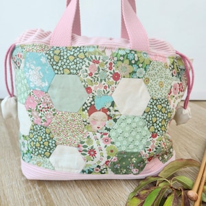 Peut inclure: Un sac à cordon doté d'un patchwork de pièces de tissu hexagonales aux motifs floraux variés dans des tons de vert, rose et blanc. Le sac présente une bordure en velours côtelé rose clair, des anses assorties et des cordons de serrage roses.