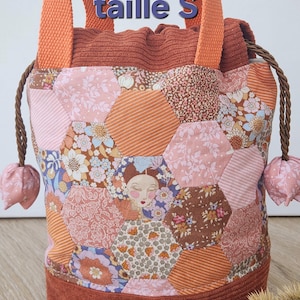 Peut inclure: Un sac seau à cordon doté d'un patchwork de panneaux en tissu hexagonal dans des tons de rose, orange et marron avec des motifs floraux et rayés. Le sac possède des anses tissées orange, une fermeture à cordon et le texte "taille S".
