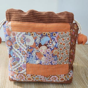 Peut inclure: Une pochette à cordon avec un motif patchwork de tissus floraux et rayés dans des tons orange, marron, violet et bleu. La partie supérieure est en velours côtelé marron avec des cordons blancs terminés par des sphères en tissu rayé.