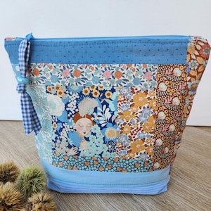 Peut inclure: Une pochette zippée en tissu patchwork avec des motifs floraux bleus, marron et orange, incluant une illustration centrale d'une personne. Les bandes supérieure et inférieure sont bleu clair uni, avec un ruban vichy bleu attaché à la fermeture.