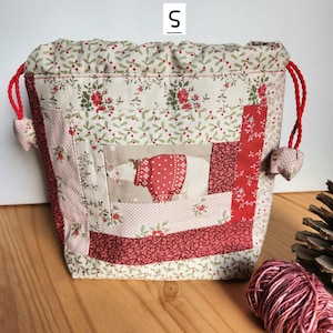 Peut inclure: Un sac à cordon fait main avec un motif patchwork. Le sac présente un ours polaire portant un pull rouge sur un panneau central. Il est orné de motifs floraux rouges et crème, avec des cordons rouges. La lettre "S" est imprimée en haut.