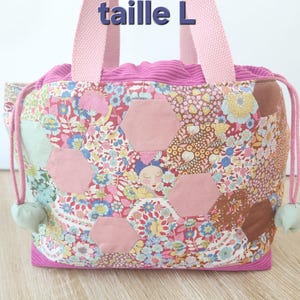 Peut inclure: Un sac à main composé d'un patchwork de tissus à motifs floraux et d'hexagones roses unis, avec une bordure en velours côtelé rose, des anses en tissu rose et une fermeture à cordon. Le texte "taille L" est inscrit en bleu au-dessus du sac.