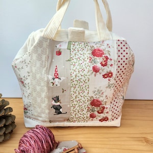 Peut inclure: Un sac fourre-tout patchwork avec des poignées et une base crème. Le sac présente des panneaux avec des motifs floraux, à pois et sur le thème des fêtes. Le sac a un design fantaisiste avec des images de chiens en tenue de fête. Une pelote de laine et des aiguilles à tricoter sont au premier plan.